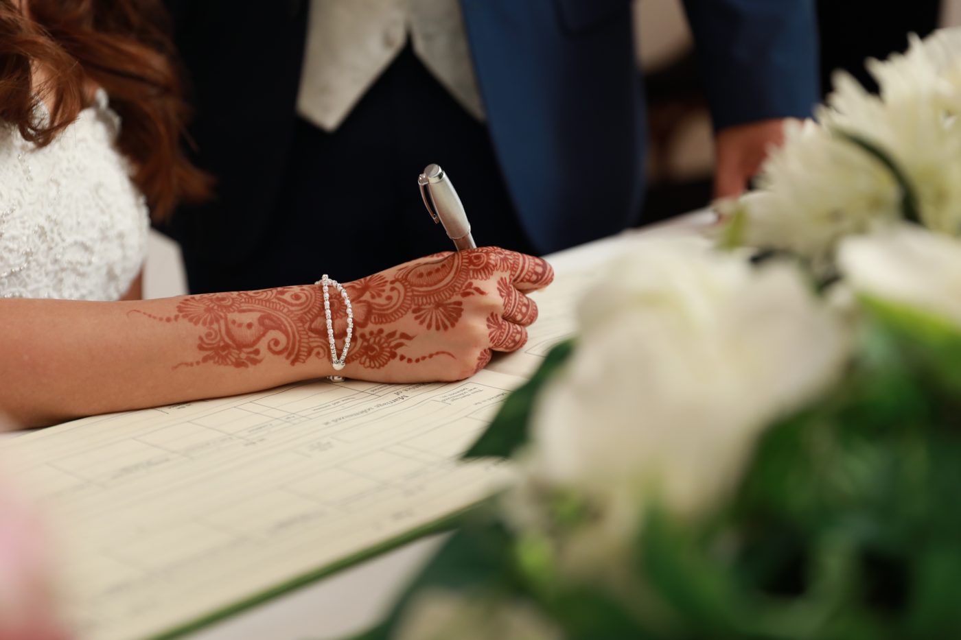 Signing the marriage register — bride's henna-decorated hand resting on the registry book, groom standing alongside, white bouquet in the foreground